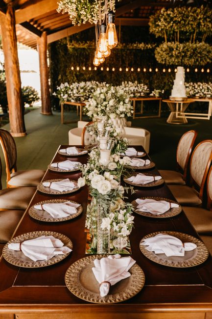 A long wooden dining table set with elegant place settings, featuring round, textured gold chargers topped with neatly folded white napkins. The table is decorated with an abundant floral arrangement 