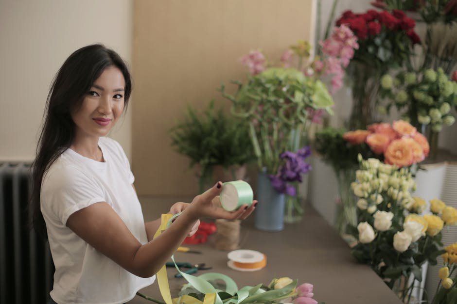A young woman with long dark hair, wearing a white t-shirt, is smiling and holding a roll of light green floral tape in her right hand, while her left hand supports a partially wrapped bouquet. The fl