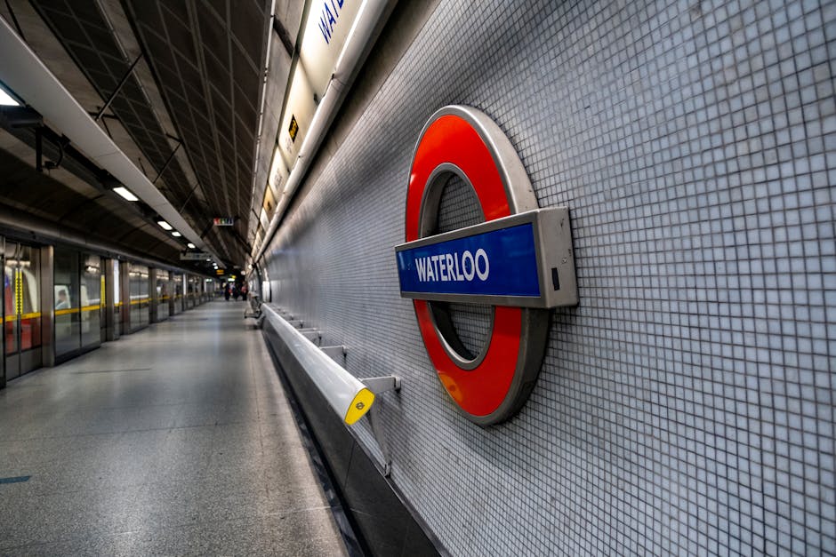 An underground platform at Waterloo station showing a tiled wall with a circular London Underground sign in red and blue with the station name 'Waterloo' displayed on a rectangular blue sign. The plat
