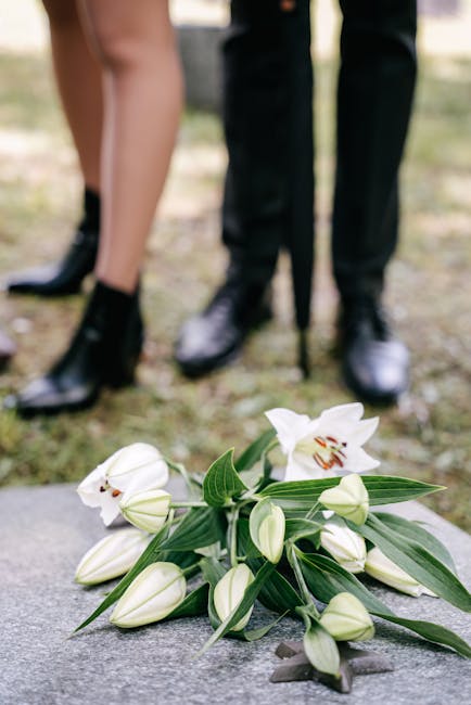 A close-up of a floral arrangement consisting of white lilies with open blooms showing reddish-brown stamens and elongated white buds, complemented by long, pointed green leaves. The bouquet is laid o